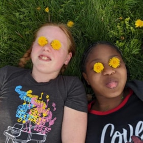 Two Girls on the Run participants lay in the grass smiling with flowers on their eyes.